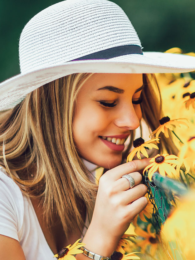 A young woman wearing a straw hat and sunglasses, smiling while standing amidst yellow flowers, seemingly enjoying the fragrance.